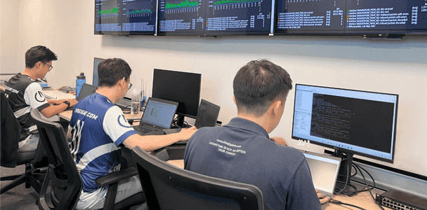 Three people in a network operations center on laptops and a desktop showing code. Multiple screens display data above them, suggesting a focused, collaborative atmosphere.