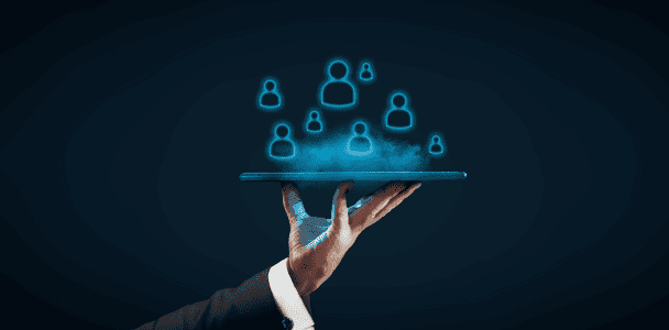 A hand in a suit holds a tablet with glowing blue icons of people floating above it, symbolizing digital connection and networking. Dark background.