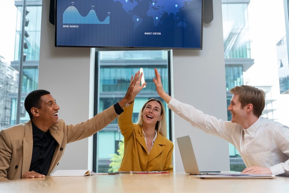 Three colleagues smiling and high-fiving around a conference table, with a laptop open.