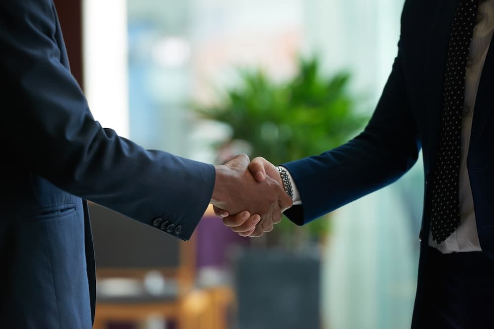 Two individuals in dark suits shake hands, symbolizing a formal agreement or partnership.