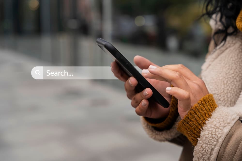 A person in a tan coat uses a smartphone outdoors. A translucent search bar with the word 'Search...' overlays the image.