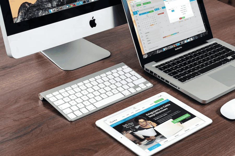A wooden desk with an Apple desktop, MacBook, iPad, and white keyboard. The iPad displays a website. The scene conveys modern work efficiency.