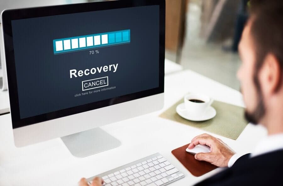 A man sits at a desk, viewing a recovery progress bar on a computer screen, showing 70% completion.