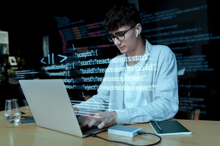 Young man wearing earphones and glasses works on a laptop with programming code overlayed.
