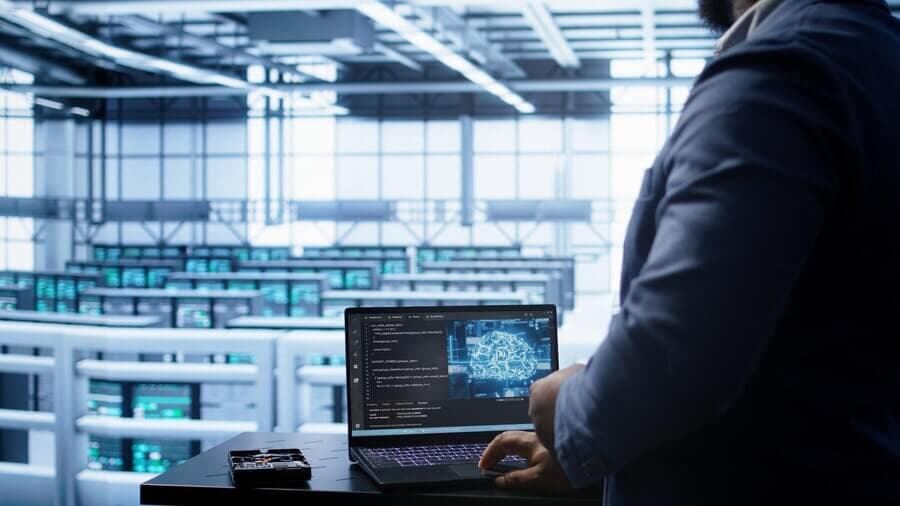 A person stands working on a laptop showing code and a digital brain diagram, set against a data center with rows of servers.