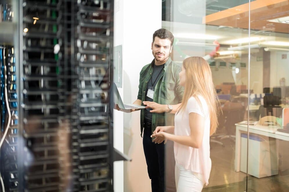 Two people are discussing with a laptop in a server room. They appear focused and engaged. Visible servers and office space set a tech ambiance.