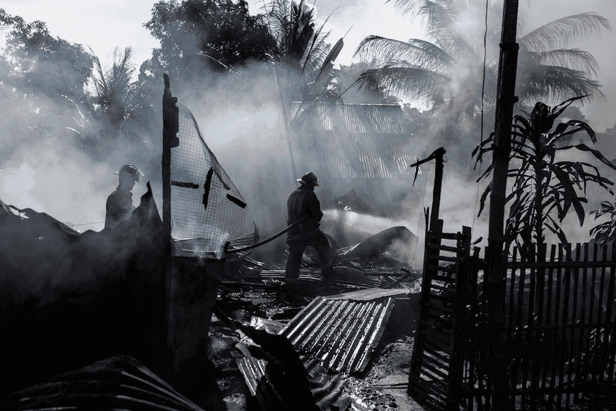 Firefighters battling a smoky blaze amid debris and ashes, silhouetted against hazy sunlight and palm trees. The scene conveys urgency and intensity.