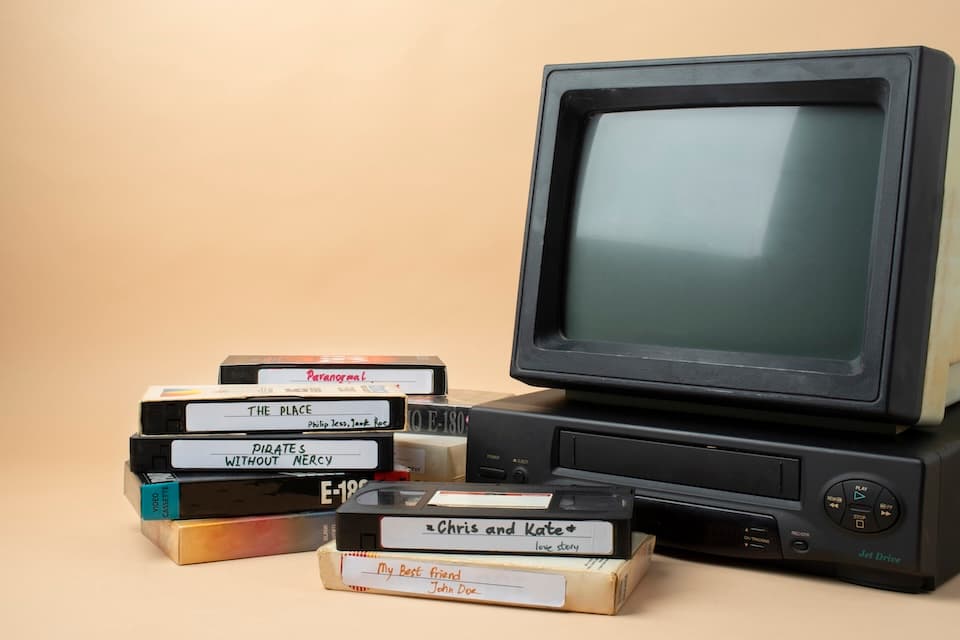 Vintage setup with a CRT TV and VHS player on the right. To the left, a stack of labeled VHS tapes.