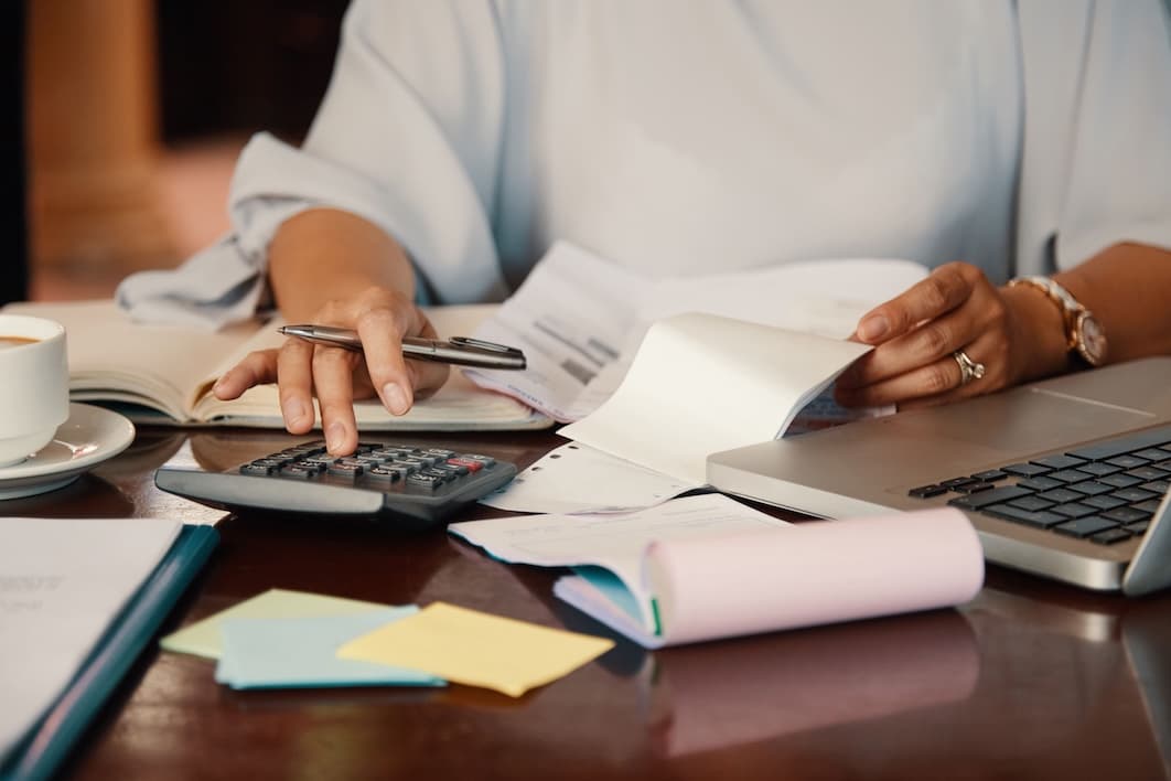 A person works at a desk with a calculator, pen, and laptop, surrounded by documents and a coffee cup, conveying focus and productivity in an office setting.