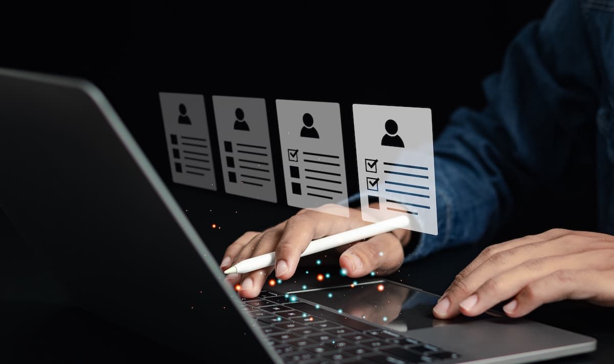 Hands use a stylus on a laptop's keyboard, interacting with floating digital profiles.