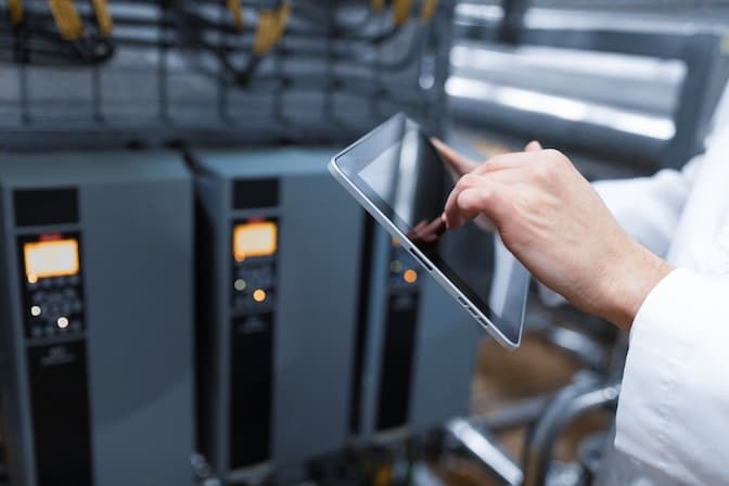 A person in a white coat uses a tablet in an industrial setting with control panels and cables. The scene conveys a sense of technology and precision.