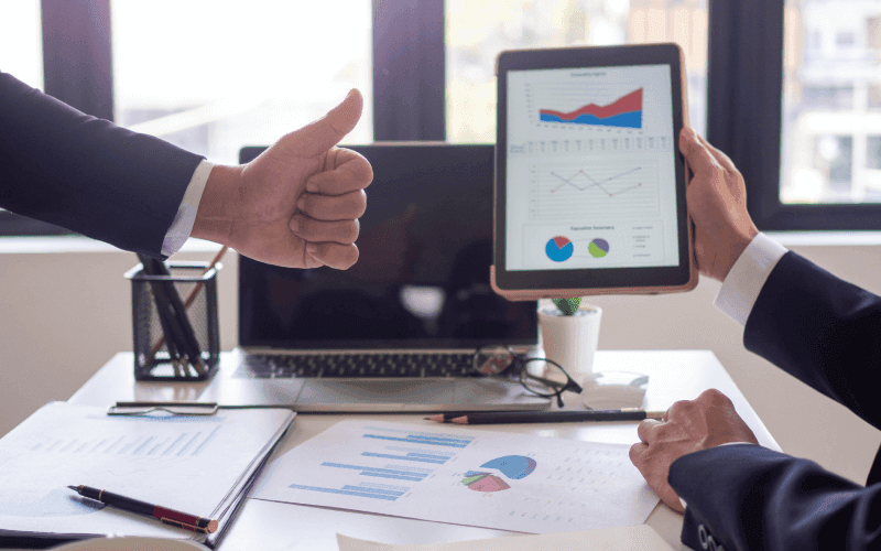 Two businesspeople in suits at a desk reviewing charts on a tablet. One shows a thumbs-up, suggesting positivity. Papers and a laptop are visible.