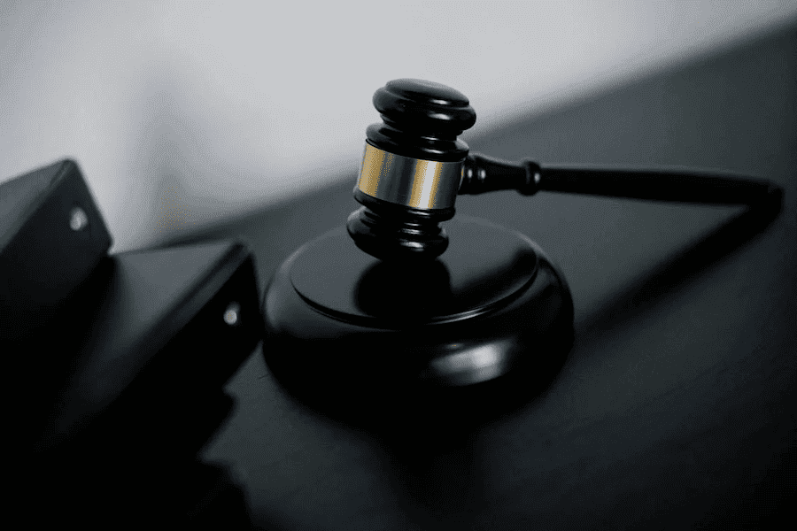 A wooden judge's gavel with a golden band rests on a sound block on a dark surface. Nearby are closed black folders, conveying a legal theme.