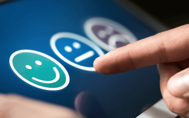 A finger hovers over three feedback icons on a touchscreen: green happy face, blue neutral face, and purple sad face, symbolizing different emotions.