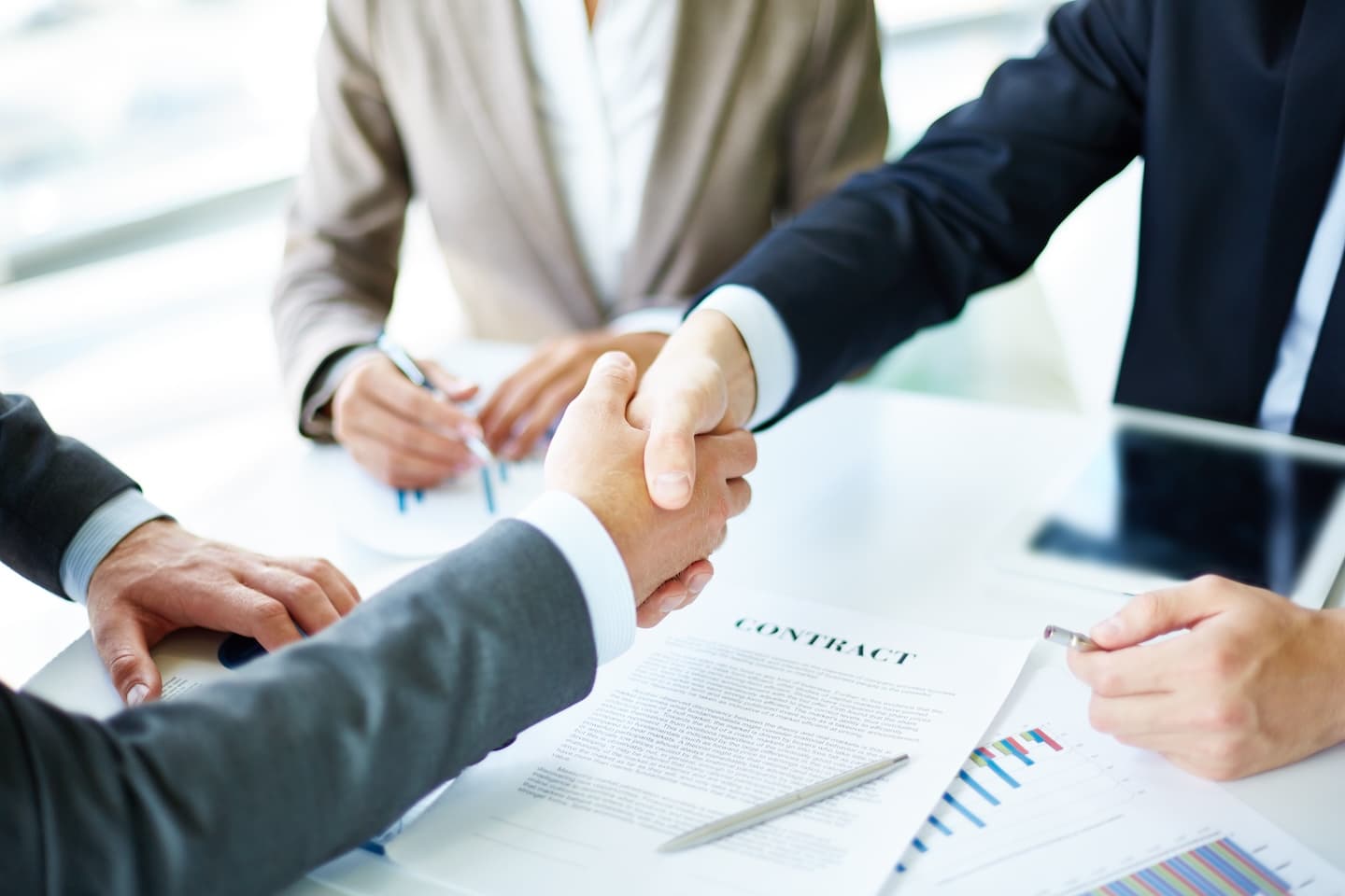 Two businesspeople in suits handshake over a contract on a table, with a pen and charts visible.